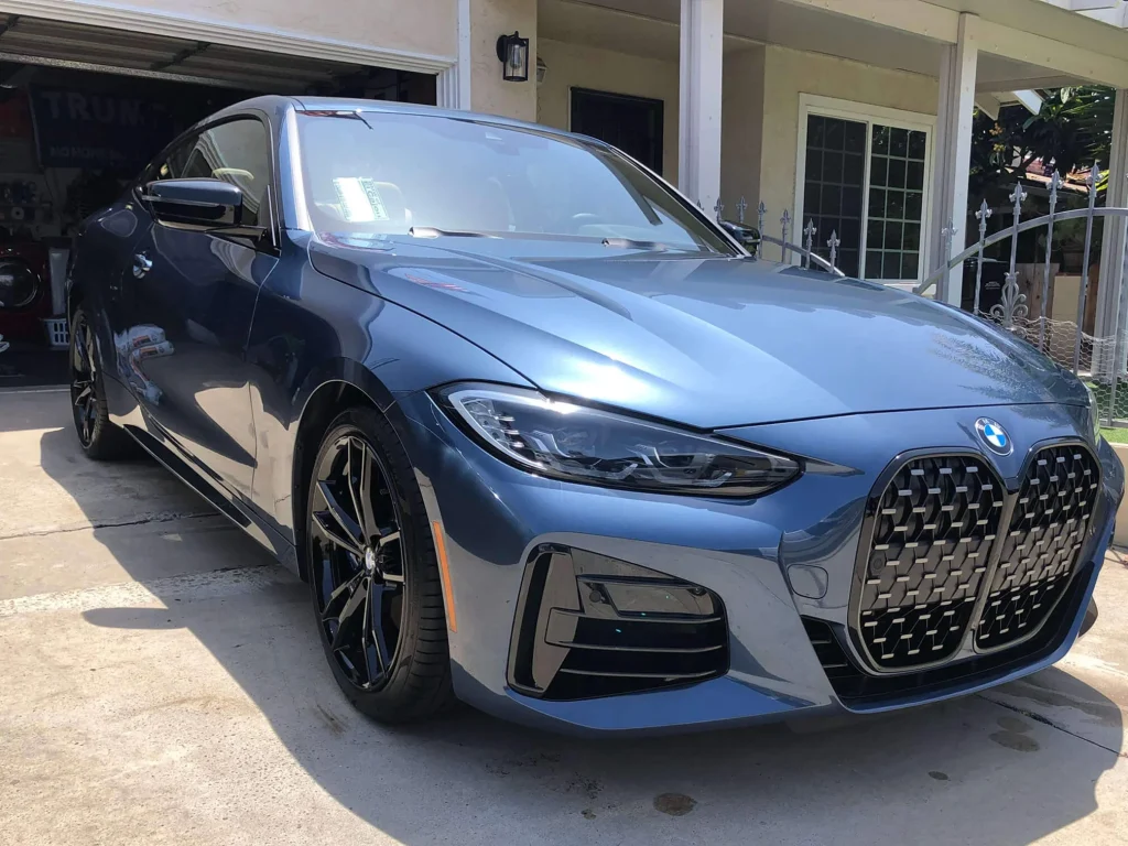 A blue BMW coupe is parked in a driveway in front of a house with a covered porch and glass doors.