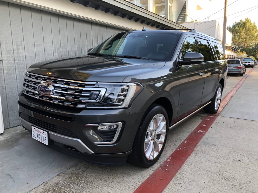 A gray Ford SUV is parked on a residential street beside a gray garage and sidewalk.