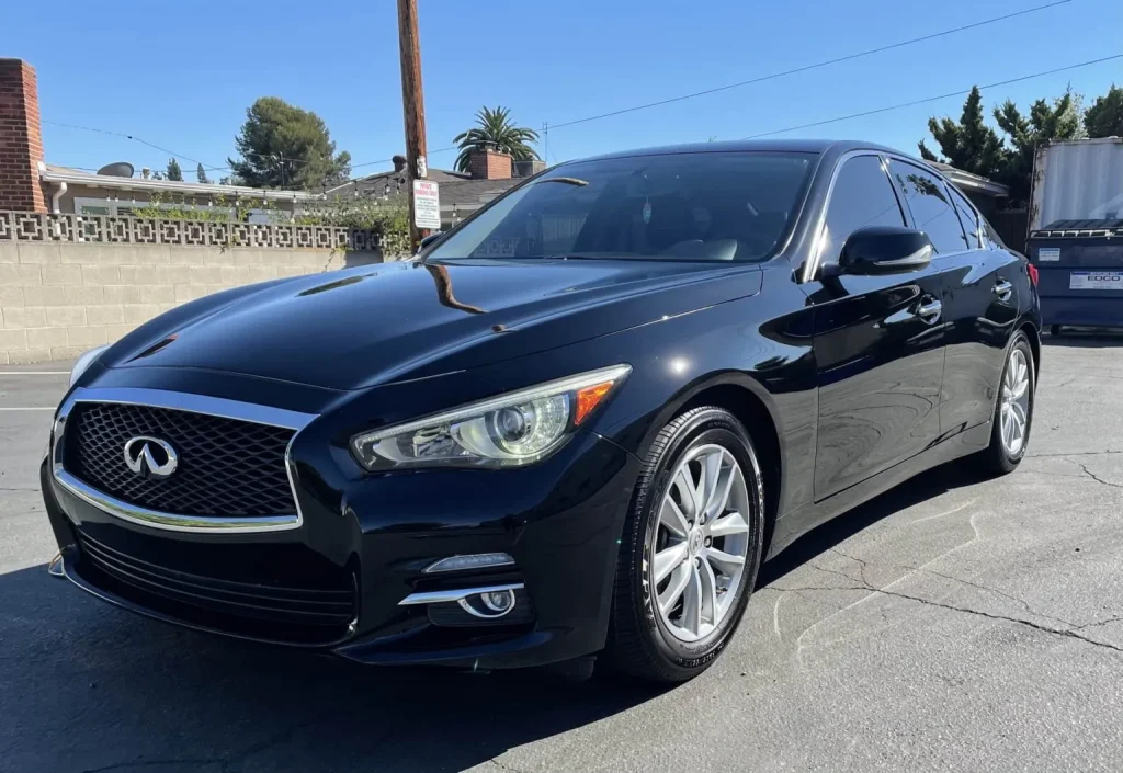 Black Infiniti sedan parked on a sunlit street near a cinder block wall and trees in the background.