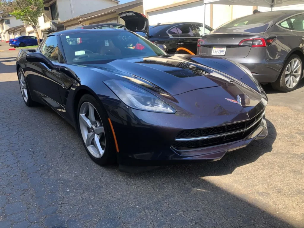 A black Chevrolet Corvette is parked on a driveway next to other vehicles under sunny conditions.